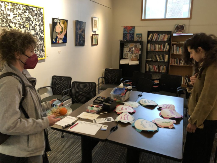 Two people standing around a table with notes and art on it. There are full bookshelves and art in the background.